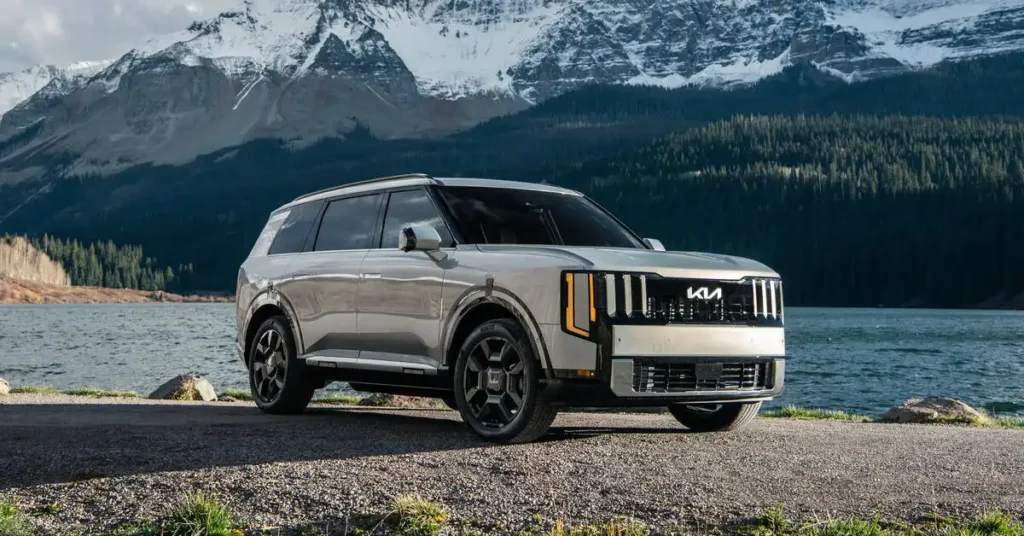 A silver Kia Telluride SUV is parked on a gravelly shore next to a calm body of water. The SUV is positioned in the foreground, angled slightly towards the viewer, with its front grille and headlights prominently displayed.