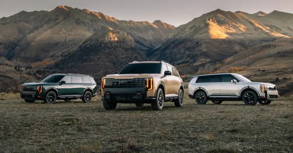 Three 2027 Kia Telluride SUVs are parked on a rocky, barren landscape with rolling hills and mountains in the background. The SUV on the left is dark green, the one in the center is a sandy gold, and the one on the right is silver.