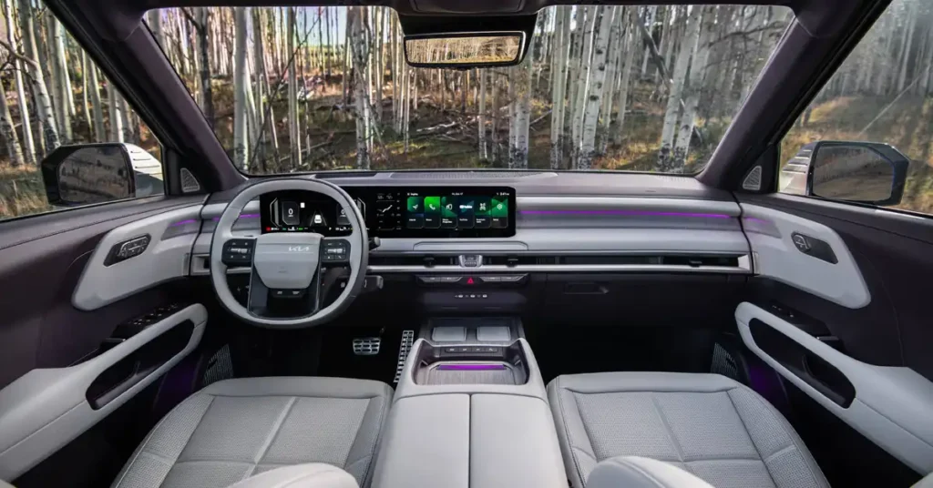 A wide-angle, eye-level shot captures the interior of a Kia Telluride SUV, looking forward through the windshield. The dashboard is dominated by a large, sleek touchscreen display showing various app icons in green. To the left, a digital instrument cluster displays a minimalist speedometer with a "0" at its center.