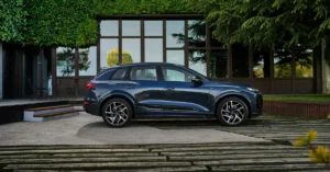 A dark blue Audi Q6 e-tron 2026 SUV is parked on a concrete driveway in front of a modern building. The building has large windows and is partially covered in green ivy.