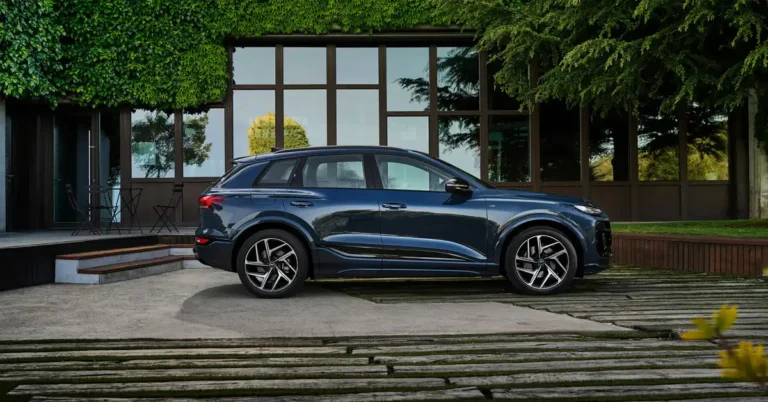 A dark blue Audi Q6 e-tron 2026 SUV is parked on a concrete driveway in front of a modern building. The building has large windows and is partially covered in green ivy.