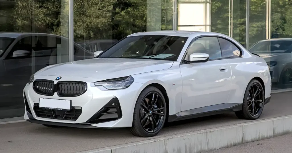 A white BMW coupe is parked in front of a modern building with large glass windows. The car is angled towards the viewer, with its front left side most prominent.