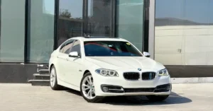 A white BMW F10 5 Series sedan is parked in front of a modern building with large glass windows. The car is positioned at a slight angle, facing the viewer.