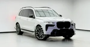 A white BMW X7 SUV is showcased from a low-angle, front-three-quarter view in a studio setting. The vehicle features a blacked-out grille and aggressive front bumper accents, contrasting with its white paint.