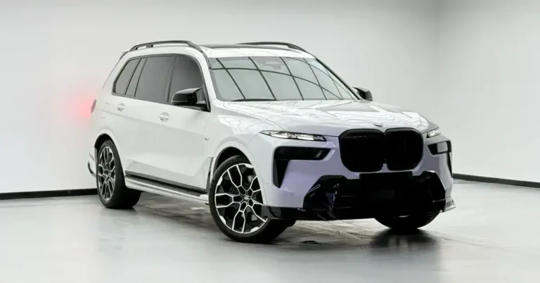 A white BMW X7 SUV is showcased from a low-angle, front-three-quarter view in a studio setting. The vehicle features a blacked-out grille and aggressive front bumper accents, contrasting with its white paint.