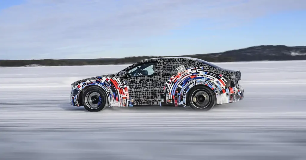A BMW M 3 car is captured in a side profile shot, moving at speed across a frozen, snow-covered landscape. The car's body is covered in a distinctive black and white grid pattern with splashes of red and blue abstract shapes, suggesting it's a prototype undergoing testing.