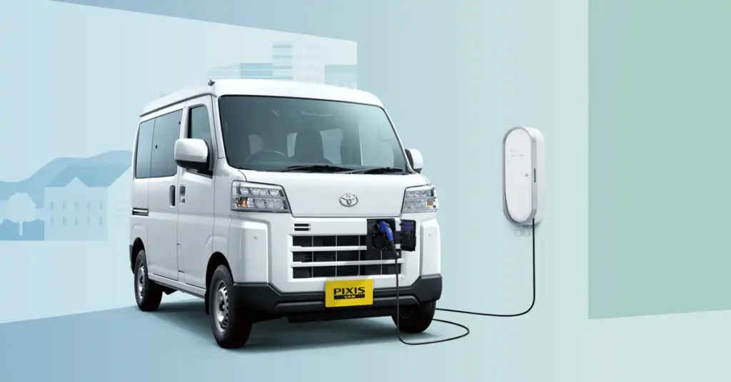 A white Toyota 2026 Pixis Van BEV, labeled "PIXIS VAN," is parked in front of a minimalist background. The van is connected to a white charging station mounted on a light blue wall. The background features a soft gradient of blue and green, with abstract, stylized representations of buildings and hills on the left side.