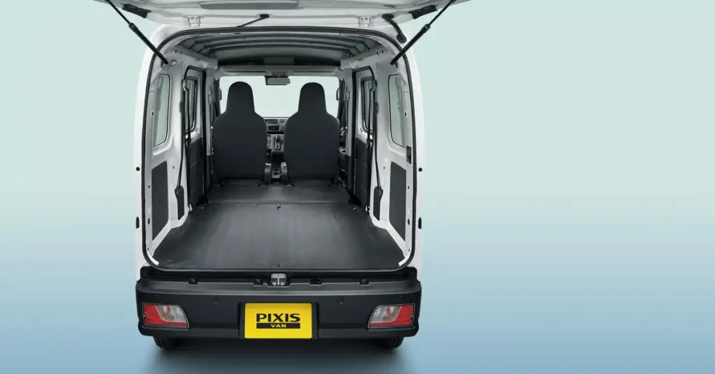 A rear view of a white Toyota Pixis Van with its back doors and trunk open, revealing the empty cargo space and two front seats. The van has a black bumper with red taillights and a yellow "PIXIS VAN" license plate. The background is a gradient of light blue to white.