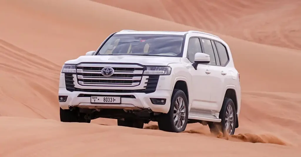 A white Toyota Land Cruiser is driving through sand dunes in the desert. The car is angled towards the viewer, with the front right side more prominent. Sand is being kicked up by the tires, indicating movement. The background consists of soft, undulating sand dunes with a warm, orange-brown hue